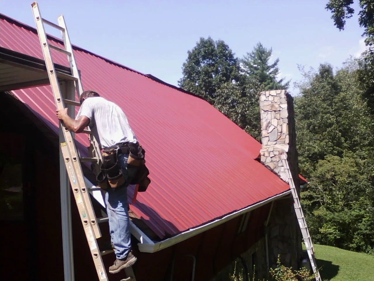 Roofer on a ladder beside a red metal roof and stone chimney — Soffit & Fascia Repair by Roofing Brothers in New Hope
